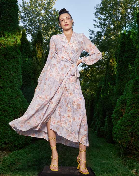 Full length image of a woman wearing a light coloured dress with a floral print, standing with her hand on her hip, surrounded by green grass and bushes.