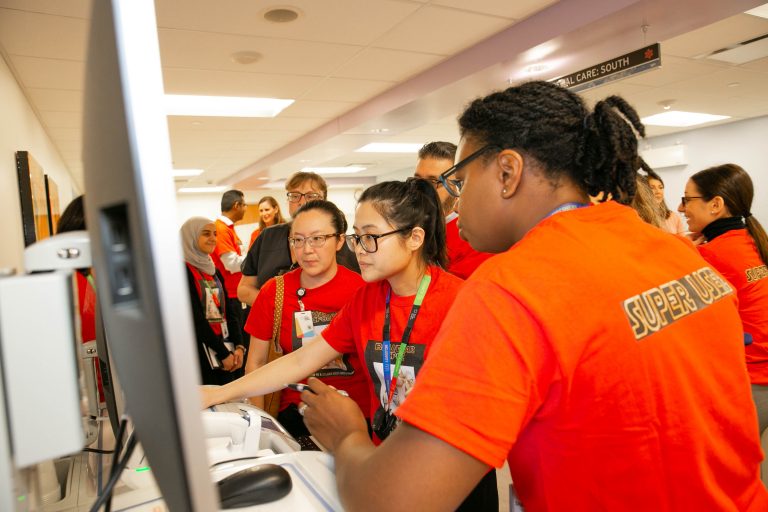 A group of health care workers on a hospital unit gathered around a computer looking at the screen. They are all wearing orange shirts