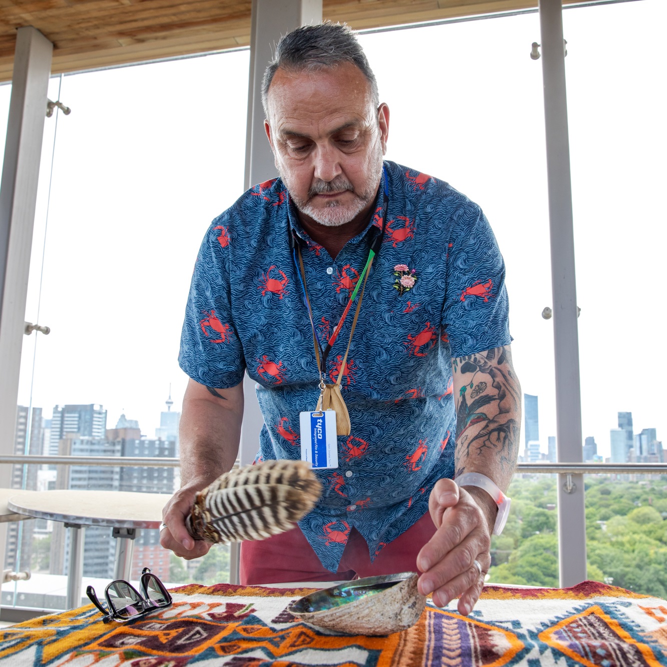 A man wearing a hospital identification badge stands over a table with a decorative cloth and a sea shell on it. He is holding a feather used to waft smoke in a smudging ceremony