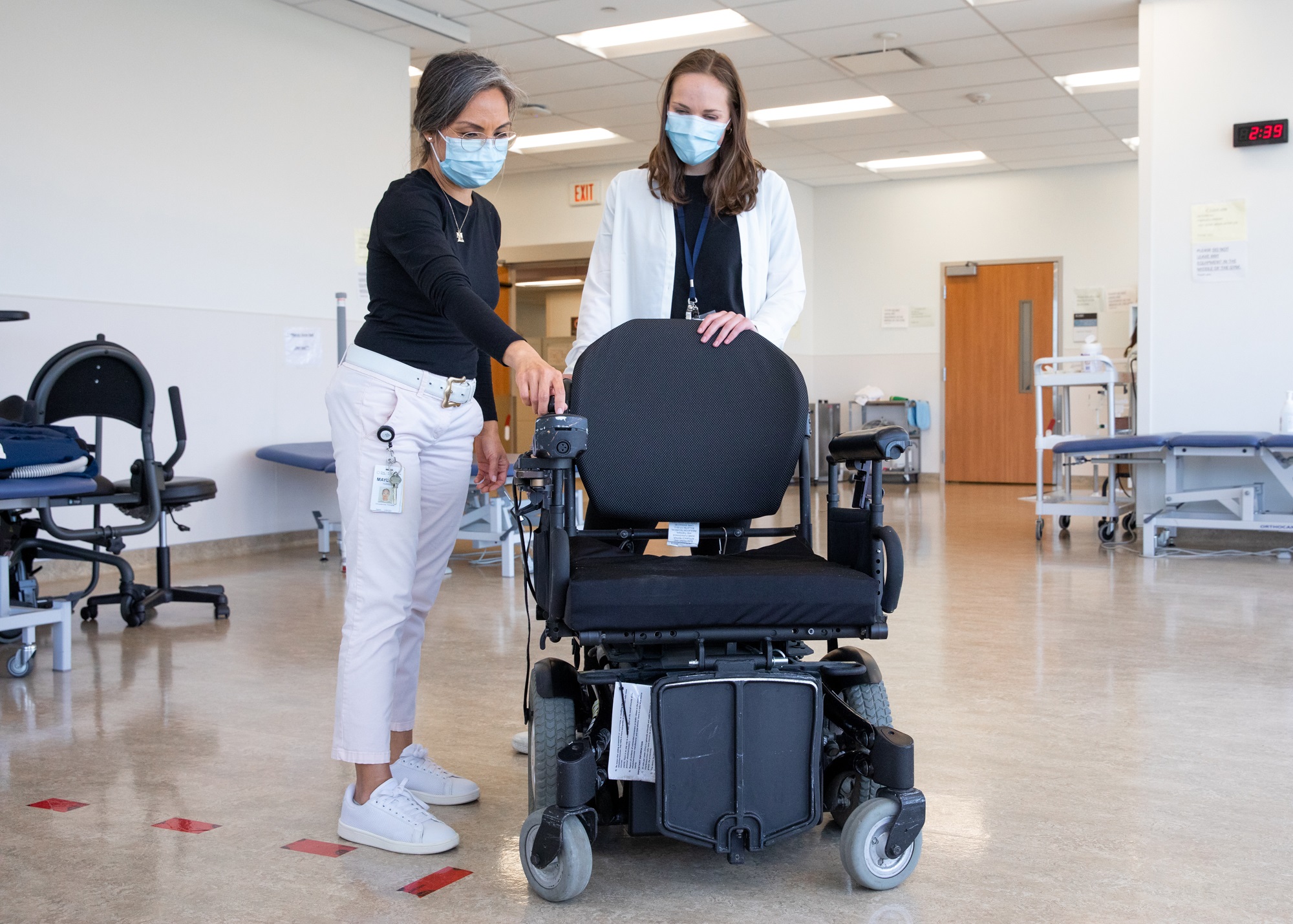 A Health care professional and a health care learner looking at a power wheelchair. The health care professional is demonstrating the controls on the armrest of the wheelchair. 