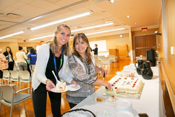 two women standing cutting cake in front of a table with coffee and plates on it