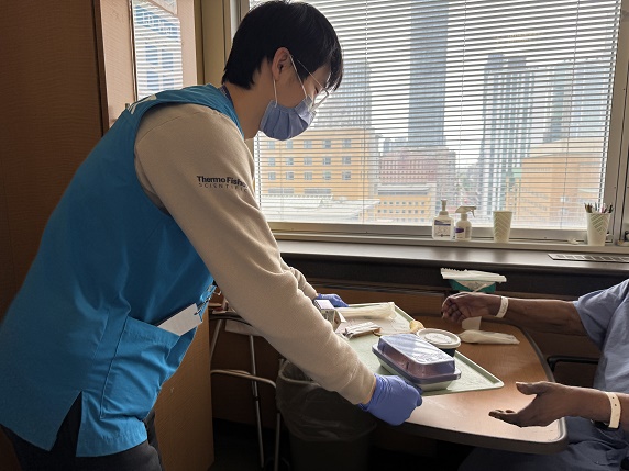 A man wearing a turquoise vest, a medical mask and gloves places a meal tray on a table for a patient in a hospital
