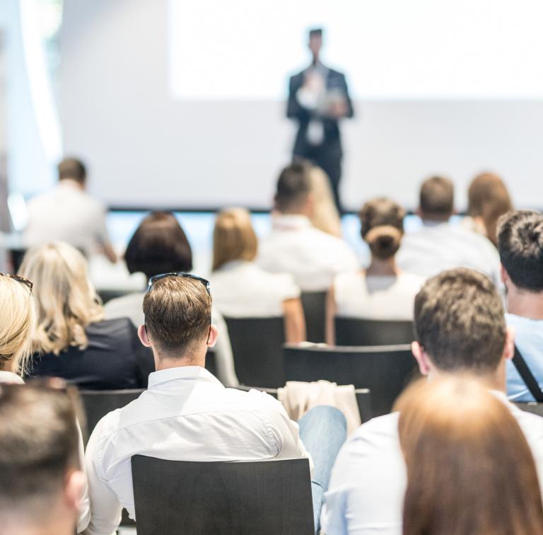 A seated audience listens to a person giving a presentation