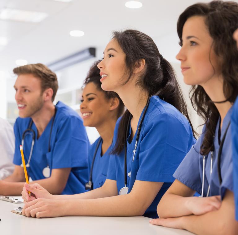 A group of people in scrubs sits at a table in a classroom