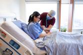 A female patient sits in a hospital bed while a physician uses a stethoscope to listen to her heart