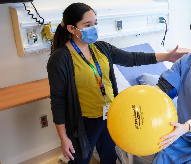 A physiotherapist looks at a patient who sits on a hospital bed and holds a yellow exercise ball