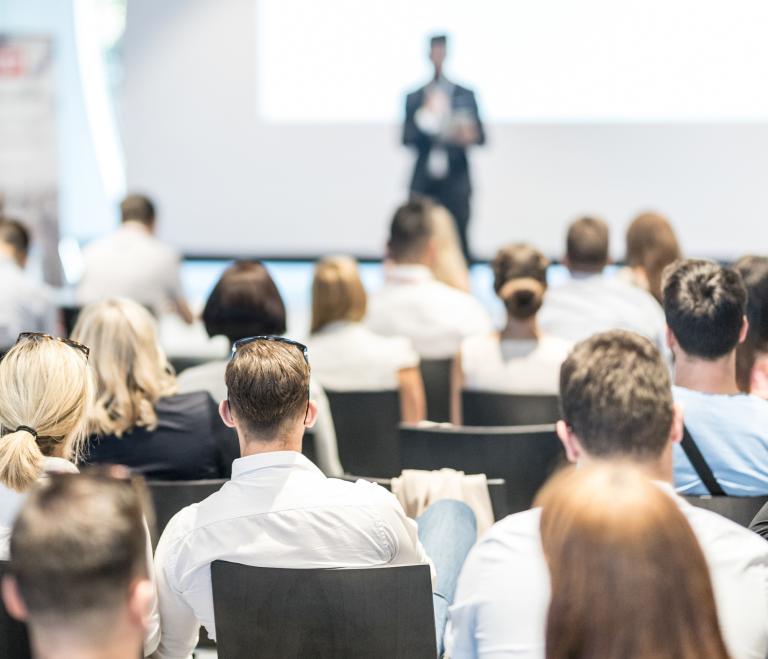 A seated audience listens to a person giving a presentation