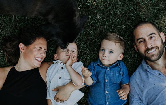 A man, a woman, their two toddler boys, laying in the grass laughing as a dog sniffs one of the toddlers