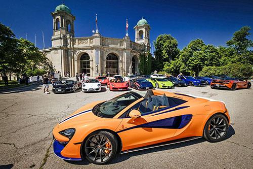 Bright orange luxury car sits in front of other luxury cars and a historic building