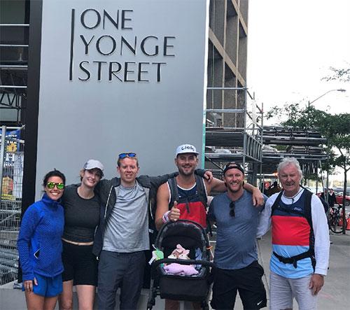 Six people, one holding a baby in a car seat, stand in front of a building that says 'one yonge street' on the side