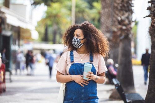 Woman stands outside, holding a coffee cup and wearing a mask