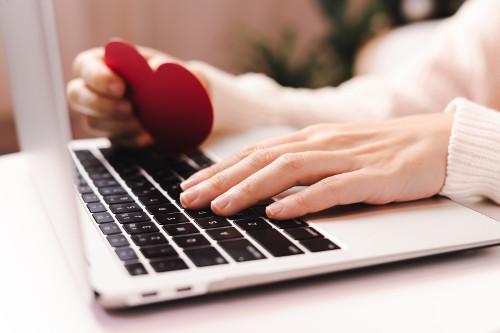 hands resting on a laptop keyboard, one is holding a red paper heart.