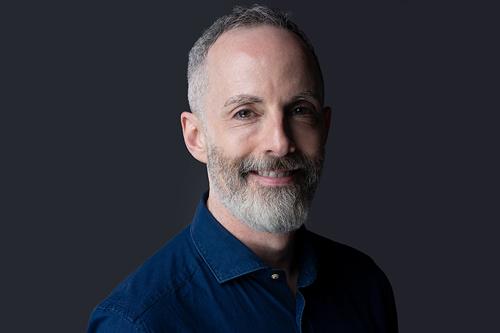 Medium close image of a smiling man with short greying hair and beard, wearing a dark blue shirt, standing against a grey background.