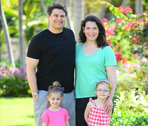 Medium long shot of a smiling family of four standing in a garden. Father wears a black shirt, mother wears a green shirt. Two young girls stand in front of them wearing pink. 