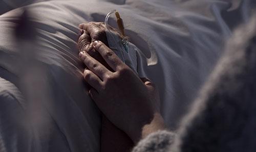 Close up shot of two hands resting on top of a white bedsheet. One hand is gently holding the other hand, which has an IV.