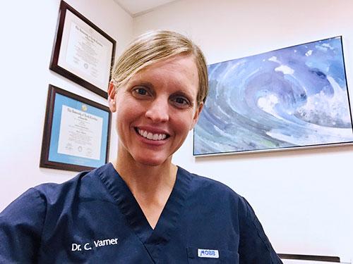 Medium close shot of a smiling blond woman, with her hair pulled back and wearing blue scrubs, sitting in an office with art and framed diplomas in the background.