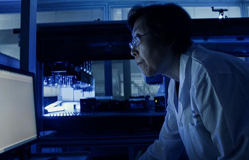 Medium close image of a woman wearing glasses and a white lab coat, standing in a dark room and leaning forward to read something on a glowing computer screen.