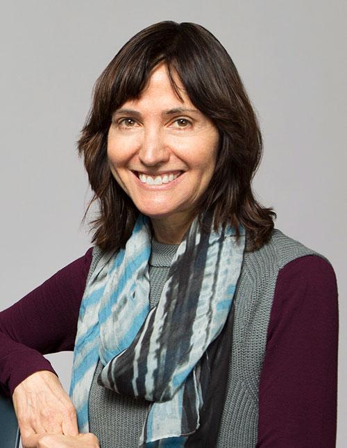 Medium close shot of a woman with shoulder length brown hair, wearing a blue and grey scarf, a knit grey vest and maroon shirt, is smiling.  