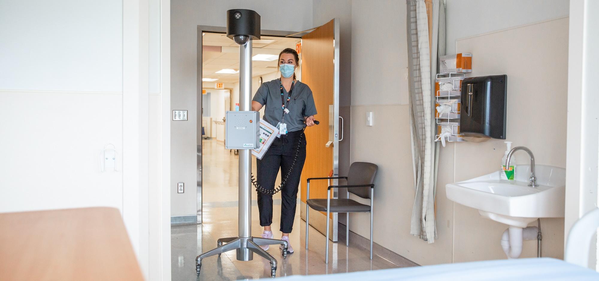 A nurse walks in to a hospital room pushing a tall metal device on wheels. 