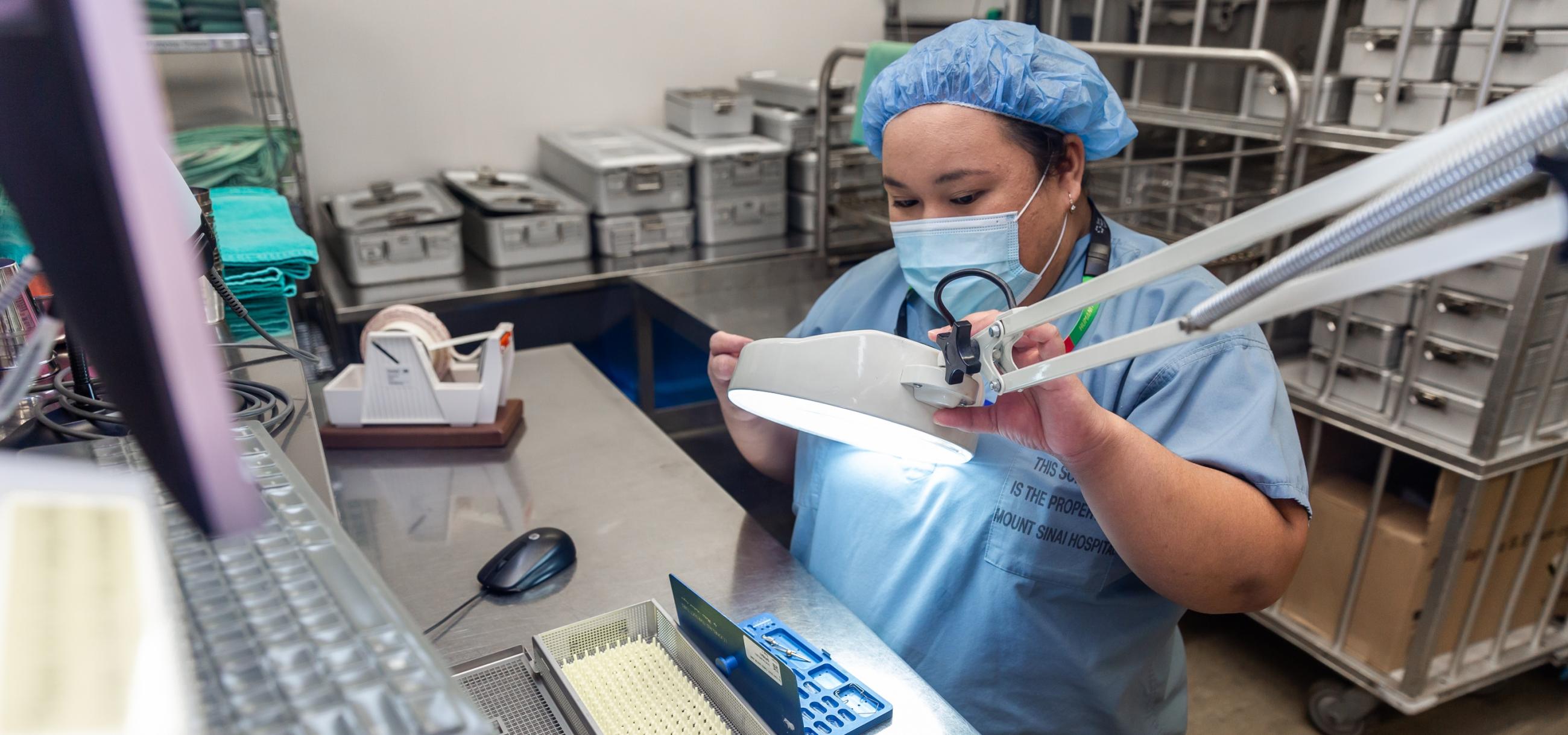 a woman wearing scrubs and a mask looks at a piece of surgical equipment through a magnifying glass