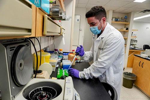 Medium close image of a man wearing a mask, a white lab coat, and purple gloves. He is transferring a substance with a pipette and there is a centrifuge in the foreground/