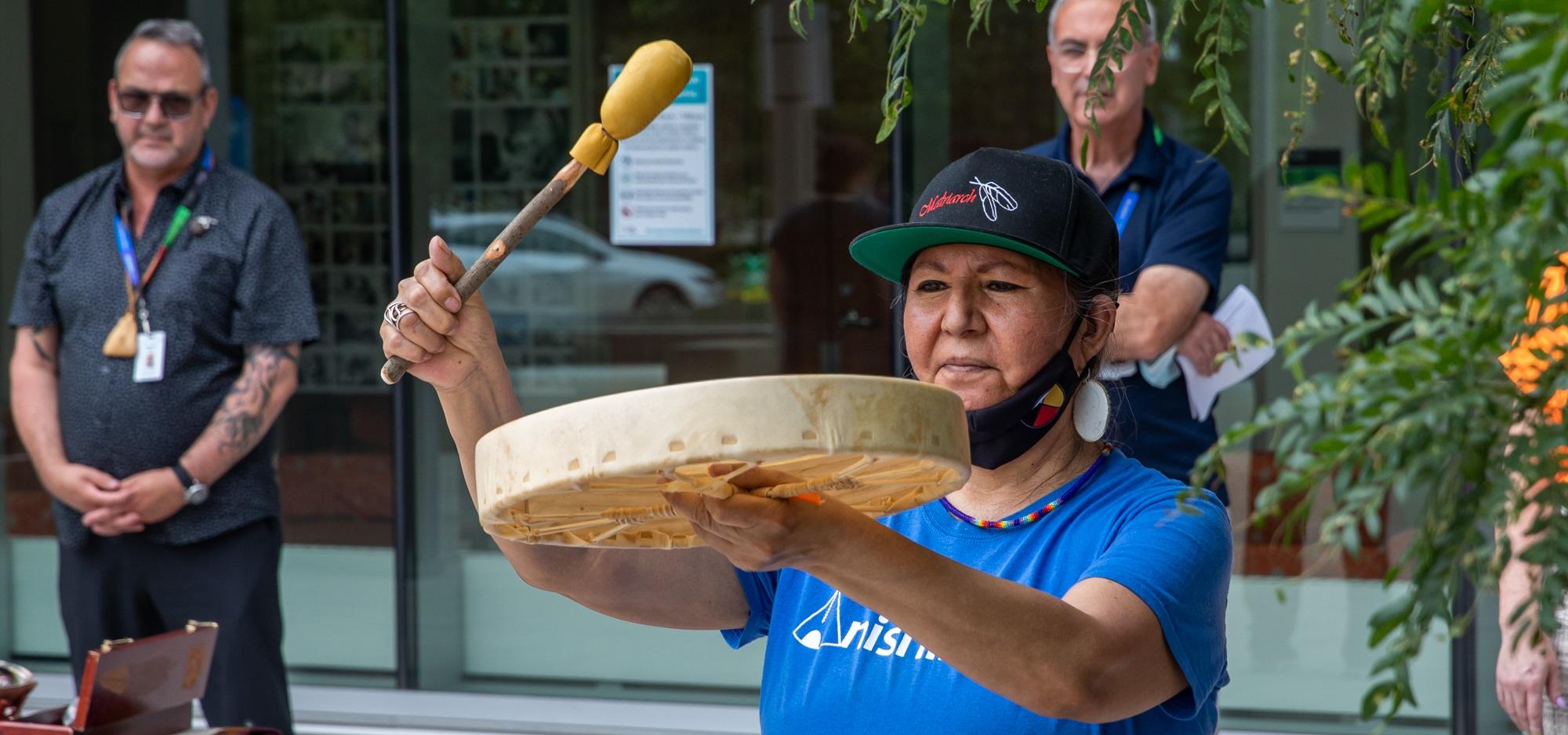 Indigenous woman plays the drum and two men are standing in the background