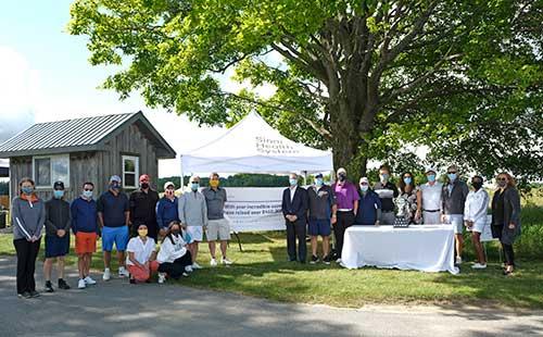 Group of people stand outside, next to a table with a trophy. 