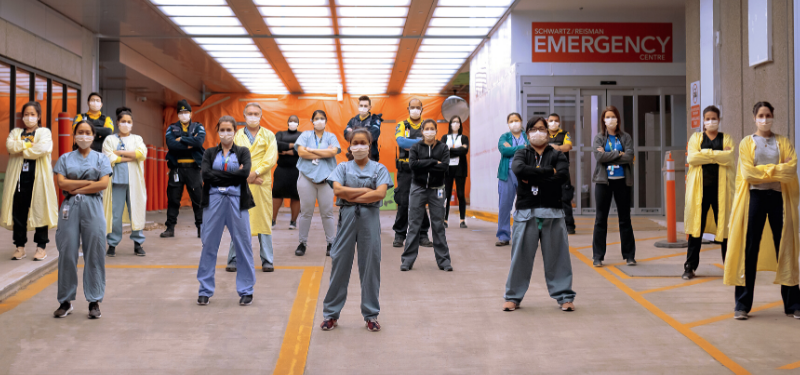 A large group of health-care workers standing outside of the Emergency department with their arms crossed. 