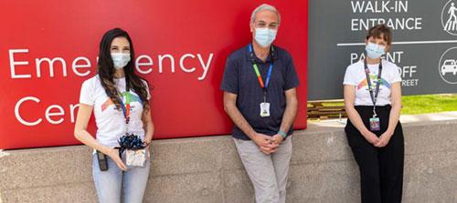 Three people (two women and one man) stand in front of a red 'Emergency Centre' sign. They each wear a mask and a coloured lanyard.