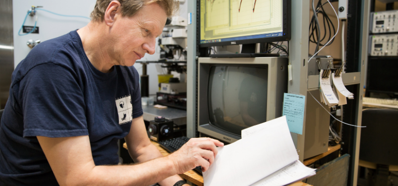 Graham Collingridge reads a printed document while sitting in front of a computer monitor.