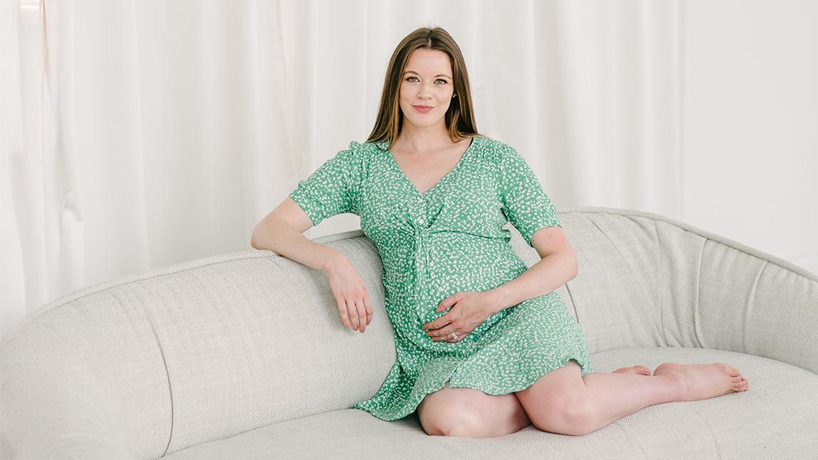 Joanna Nicholson sitting on a grey couch in a green dress. 