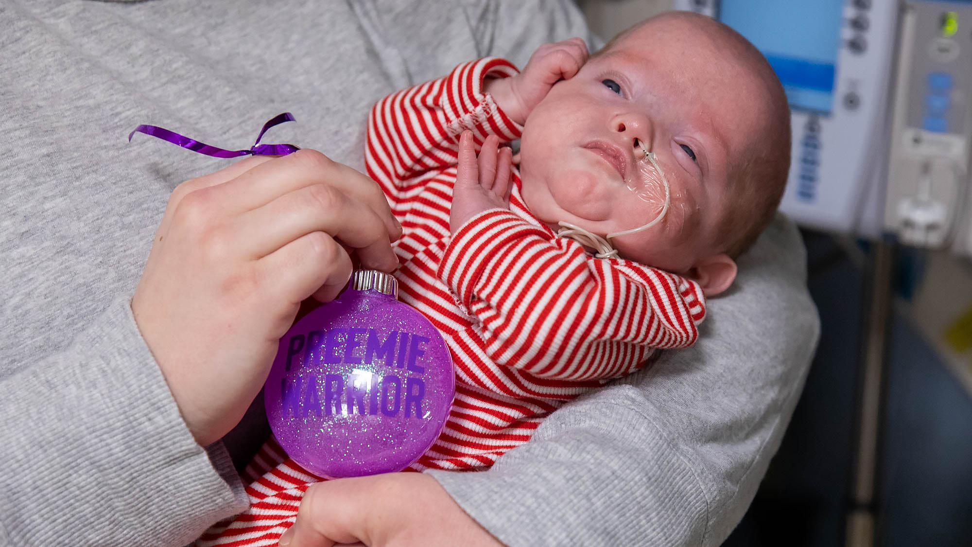 Infant in the NICU held in their mother’s arms beside a hand-made ornament. Caption: This holiday season, families in our NICU received hand-made, donated ornaments.