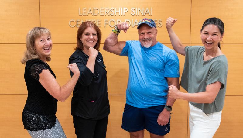 Four people pose in front of a wall that reads "Leadership Sinai Centre for Diabetes". A muscular man in a blue shirt and shorts flexes his arm, beside three smiling women - one in scrubs, one in a black lace top, and one in a light green top - who are also playfully flexing their arms.