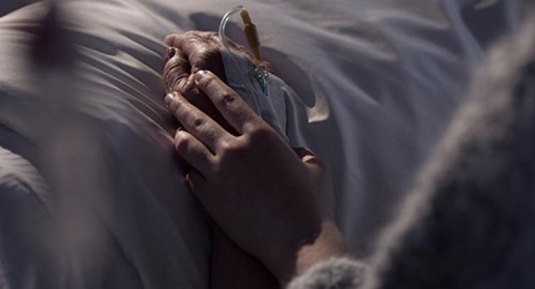 Close up shot of two hands resting on top of a white bedsheet. One hand is gently holding the other hand, which has an IV.