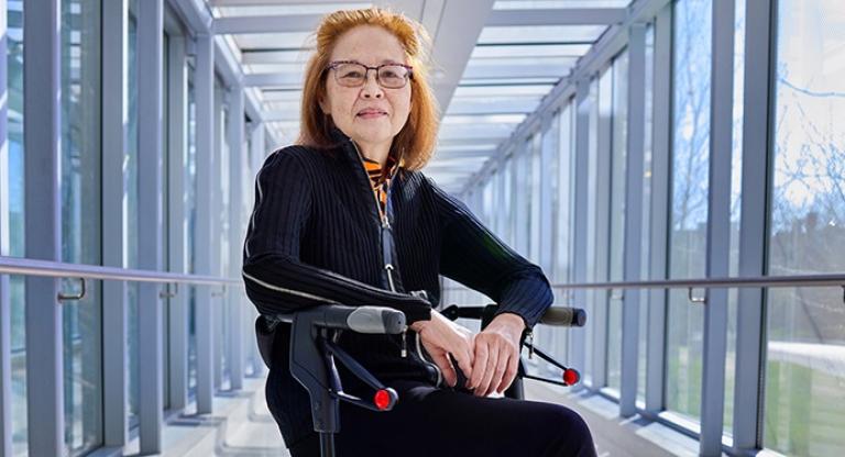 A woman sits on a rollator mobility device. She is surrounded on both sides by the windows of an indoor walkway. She is looking at the camera smiling with her hands clasped together. She has auburn hair, glasses and is wearing a black.