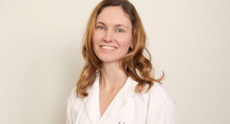 Medium close shot of a woman with shoulder length blond hair, wearing a white lab coat and smiling. 