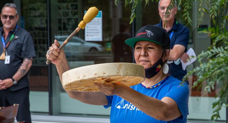 Indigenous woman plays the drum and two men are standing in the background