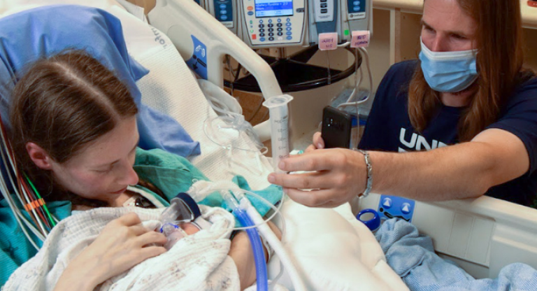 A woman and her baby in bed while a loved one is by her side