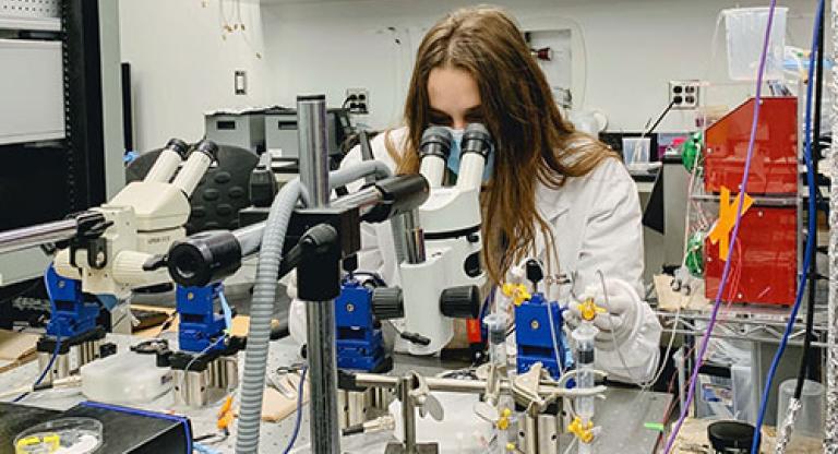 Medium close image of young woman with long hair sitting at a medical work station surrounded by equipment, looking into a microscope. 