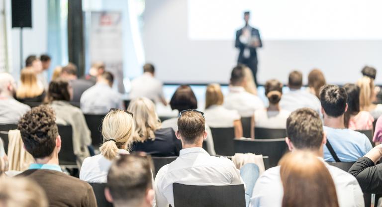 A seated audience listens to a person giving a presentation