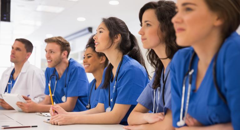 A group of people in scrubs sits at a table in a classroom