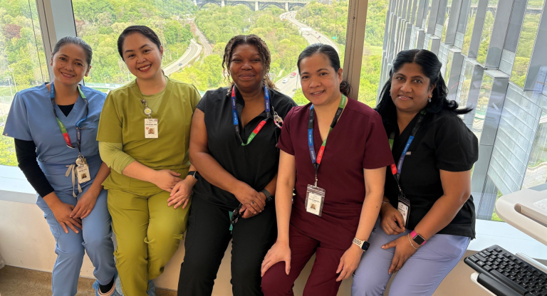 A group of four hospital employees wearing scrubs. They are sitting in a row in front of a window with a view of a green leafy valley behind them. They are looking at the camera and smiling