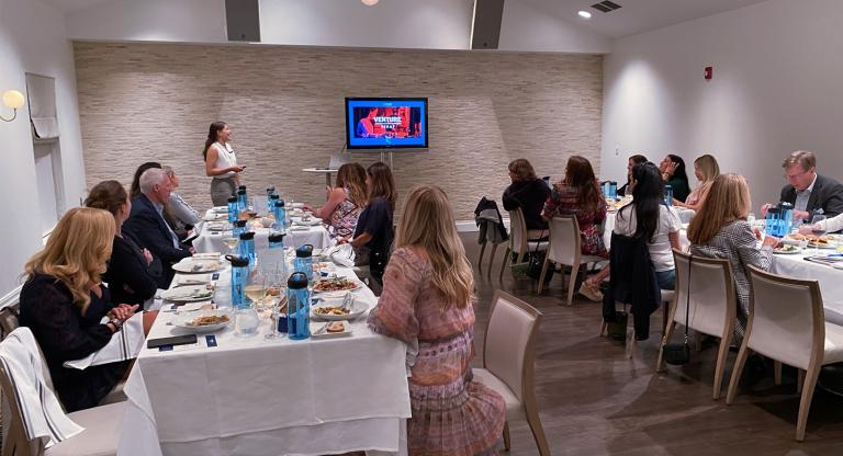 Group of people sitting at table watching a woman give a lecture