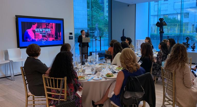 Group of people sitting at table watching a woman give a lecture